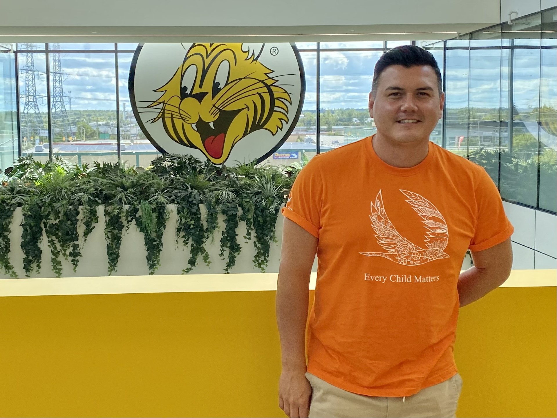 Patrick Hunter standing in front of Giant Tiger head office logo, wearing an orange T-shirt he designed, for his campaign with Giant Tiger and Indspire