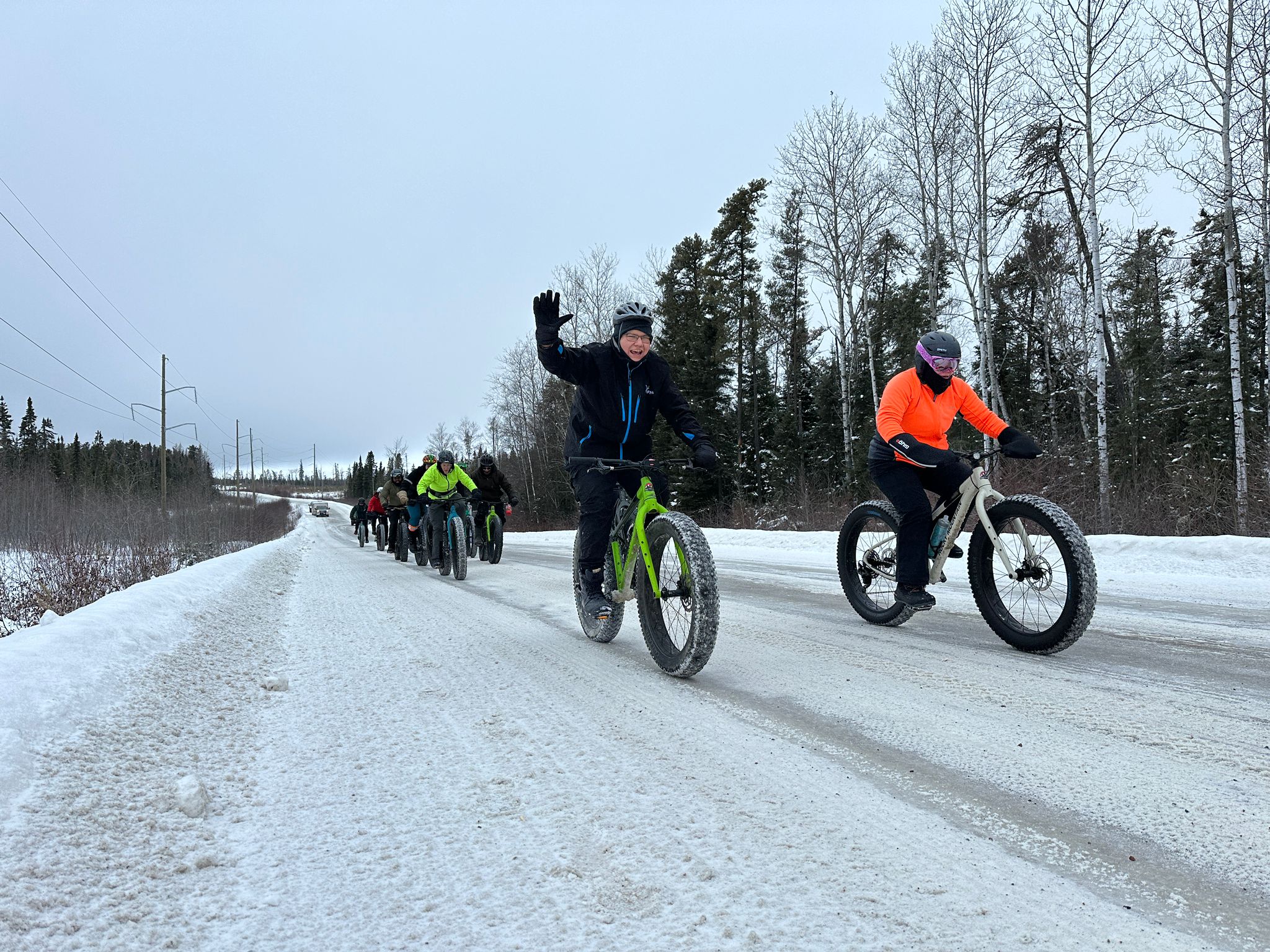 Bike riders on fat bikes waving at the camera on a snowy trail in the forest.